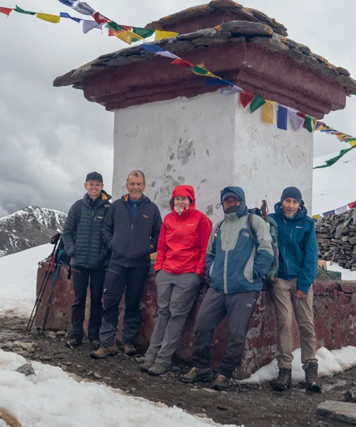 Five hikers stand in the snow by a traditional stone chorten with prayer flags during their Jomolhari Base Camp Trek through the Bhutanese Himalayas.