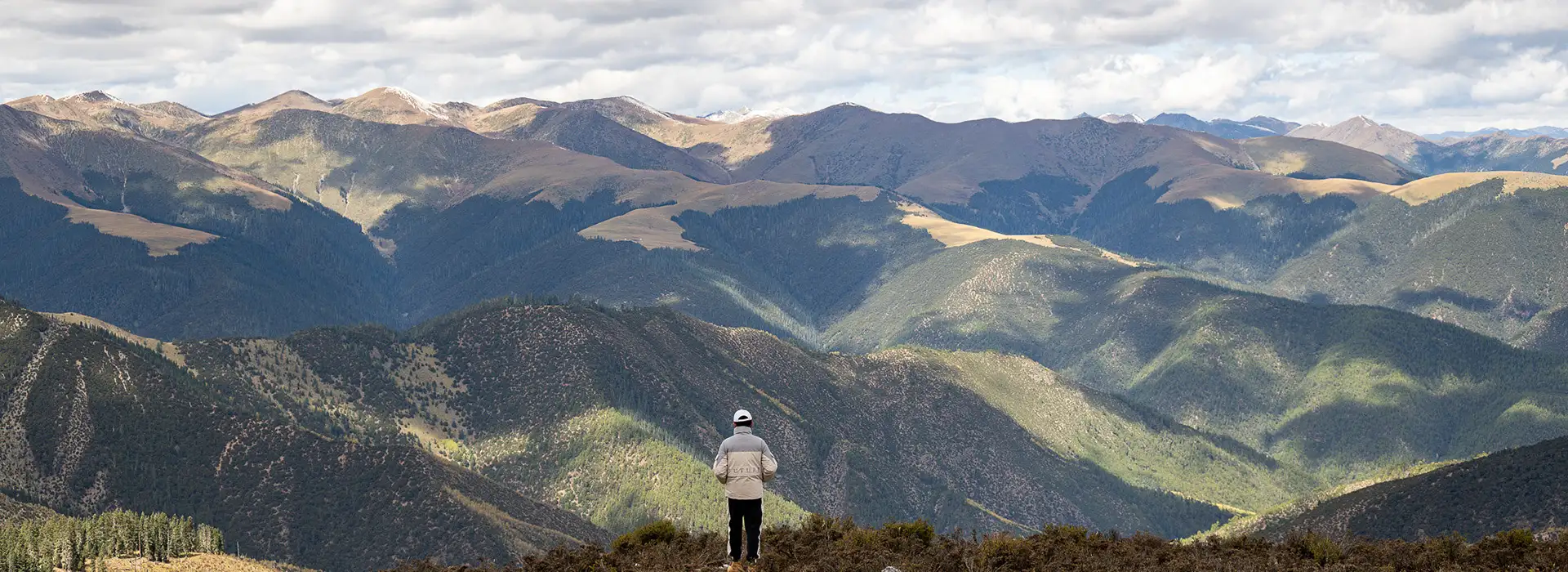 A lone traveler in a white cap stands on a mountain ridge, overlooking a vast range of deep green valleys under a cloudy sky during a Kathmandu to Lhasa Tour.
