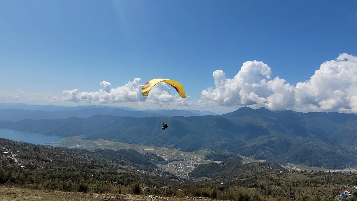 A wide panorama shows a yellow paraglider with two people soaring high above a mountain range, overlooking a valley and distant lake, perfect for an adventurous Pokhara Travel Guide.