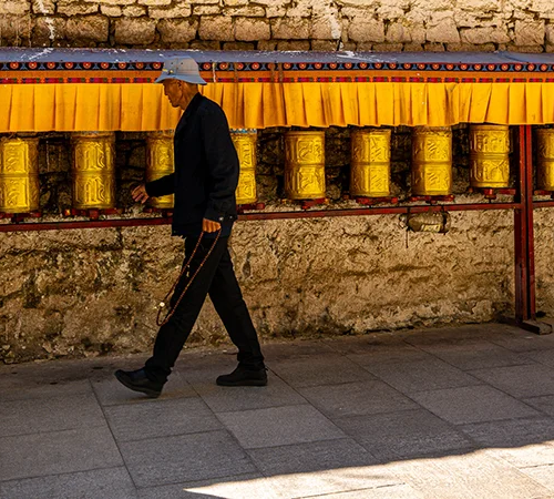 An elder pilgrim with prayer beads spins a row of golden prayer wheels along a stone wall, a spiritual scene common on a Lhasa to Kathmandu overland tour.