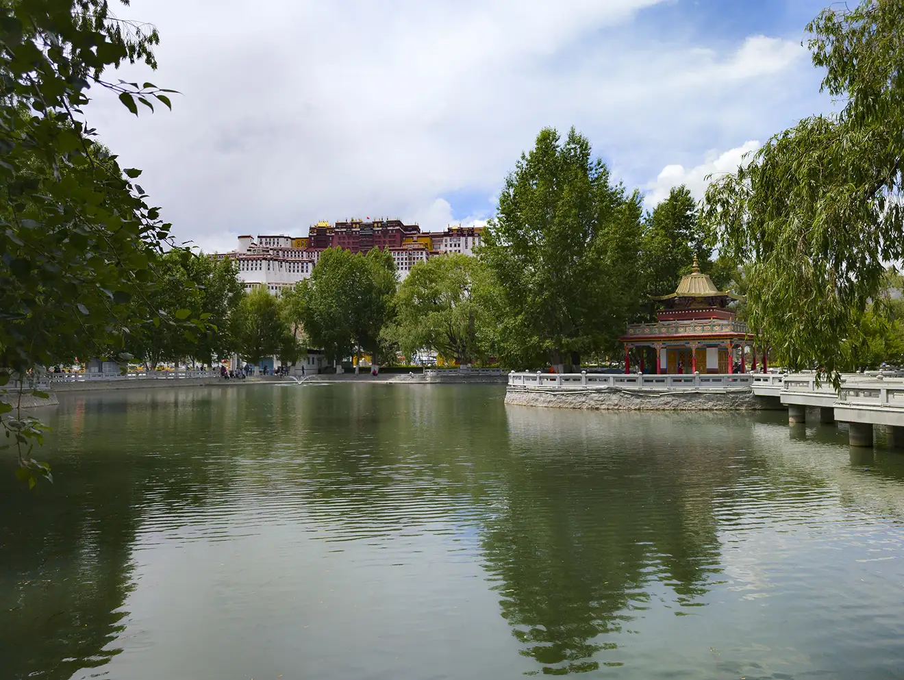 View of Potala Palace above a lakeside park in Lhasa during the Lhasa to Everest Base Camp and Namtso Lake Tour, with trees and a pavilion reflected in calm water.