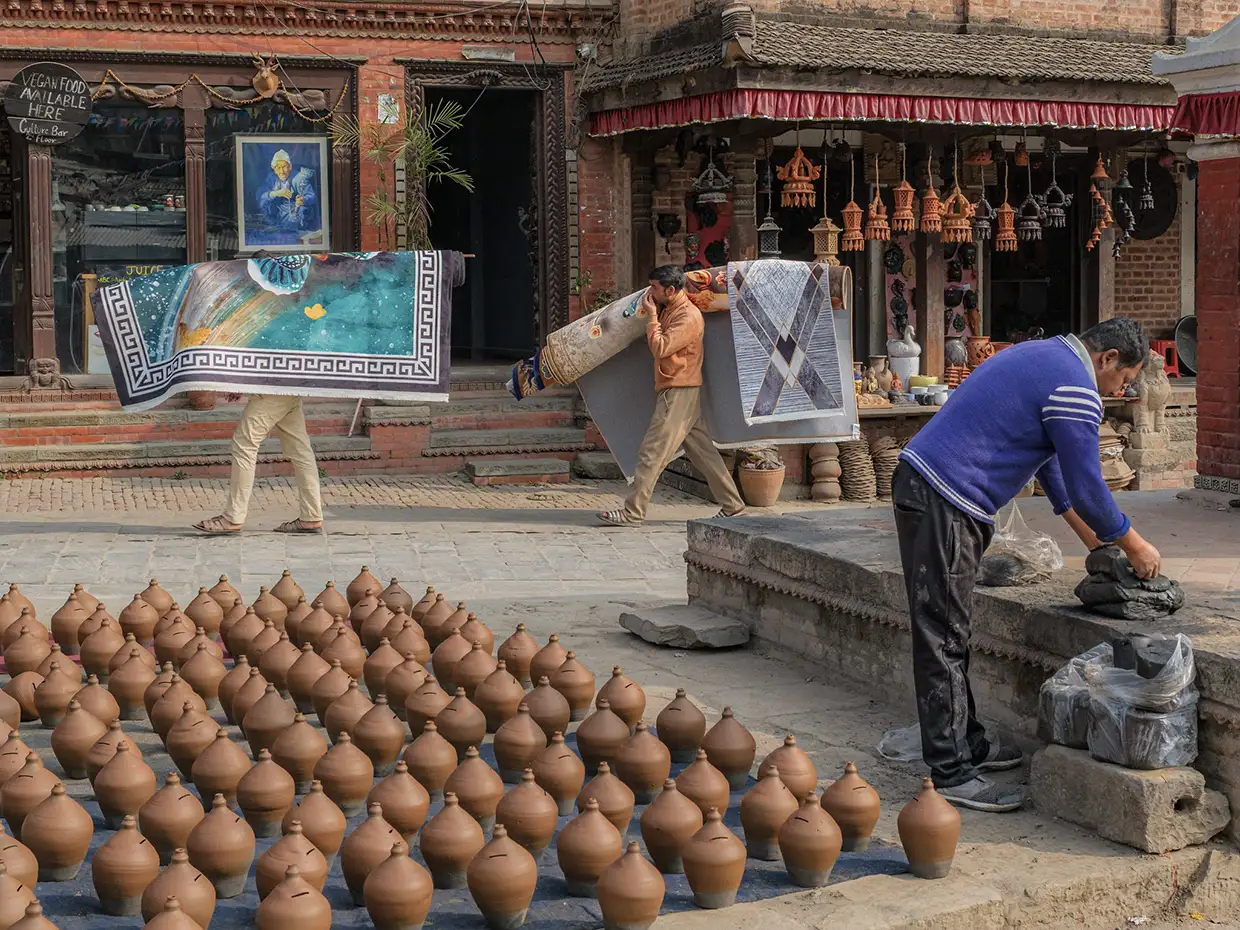 Dozens of clay pots dry in a sunny brick courtyard while local men carry carpets and work with clay, illustrating things to do in Kathmandu.