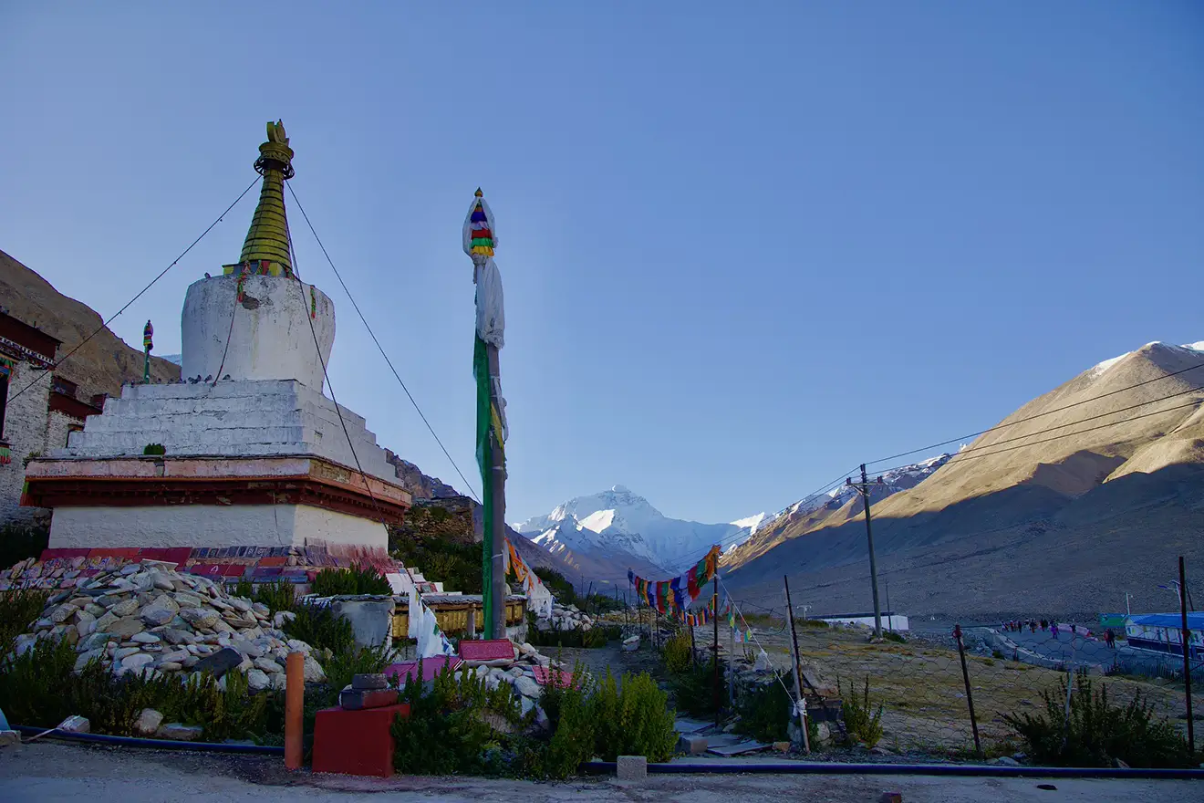 A wide shot featuring a traditional Tibetan chorten, colorful prayer flags, and the snow-capped peak of Mount Everest in the distance under a clear sky.