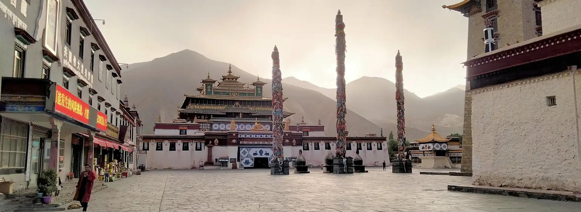Tibetan monastery courtyard at sunset, featuring the main temple with gold roof, prayer flag poles, and distant mountains. This stunning site is a highlight of our Lhasa and Shigatse Tour.