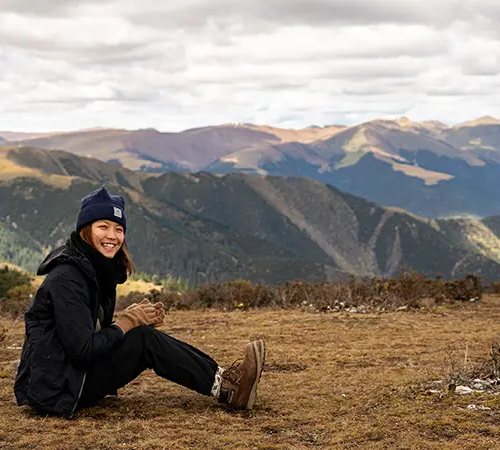 A smiling female traveler sits on a grassy plateau with rolling mountain ranges behind her. Many people plan a Tibet tour from India to experience these views.