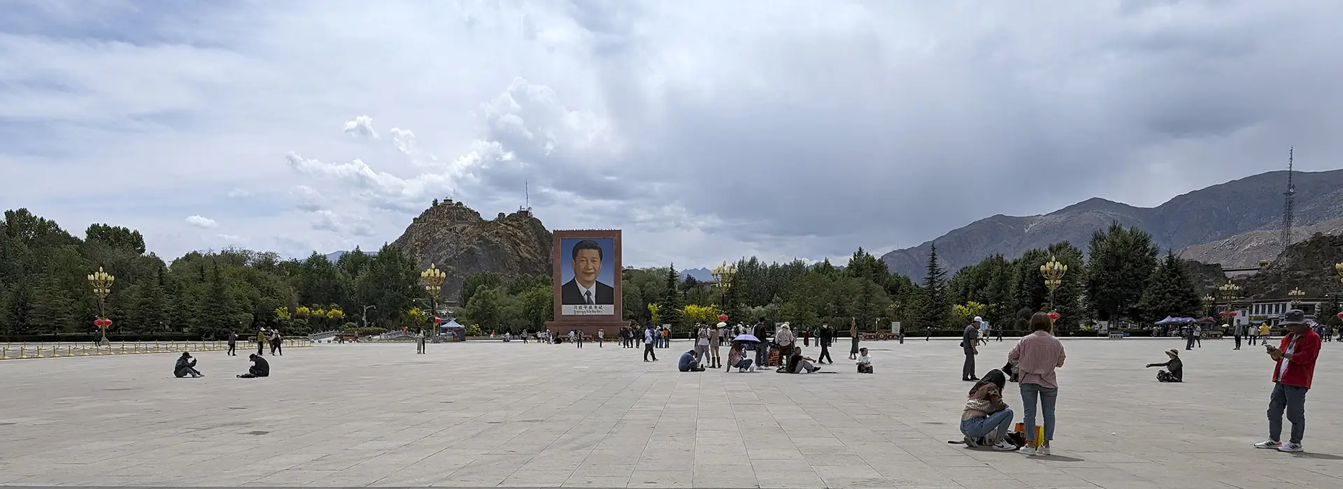 Tourists explore a large square in front of mountains with a large outdoor portrait during a Lhasa Short Tour under an overcast sky.