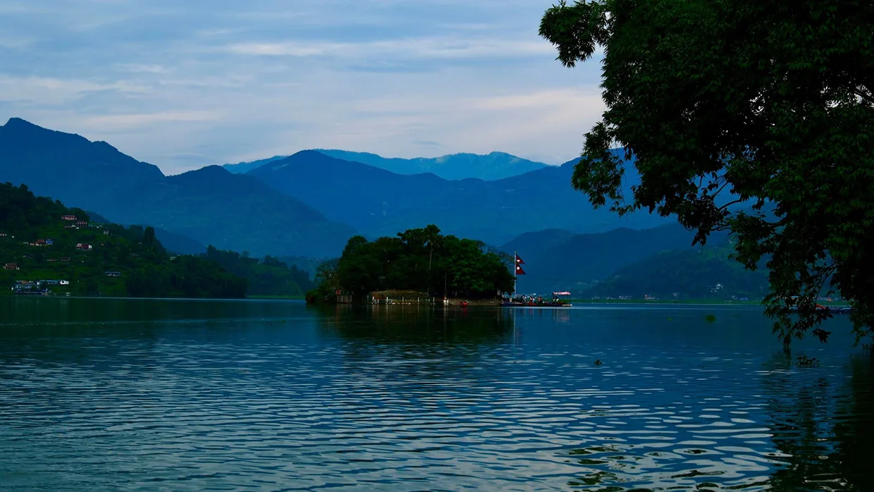 A beautiful scenic photograph of Phewa Lake in Nepal, showing the Tal Barahi Temple island and majestic mountain views from a lush shoreline, perfect for a Pokhara Travel Guide.
