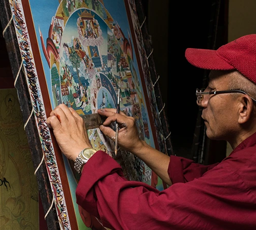A detail shot showing a skilled artist in a red shirt and cap painting a traditional thangka, highlighting the meticulous craftsmanship found when searching for things to do in Kathmandu.