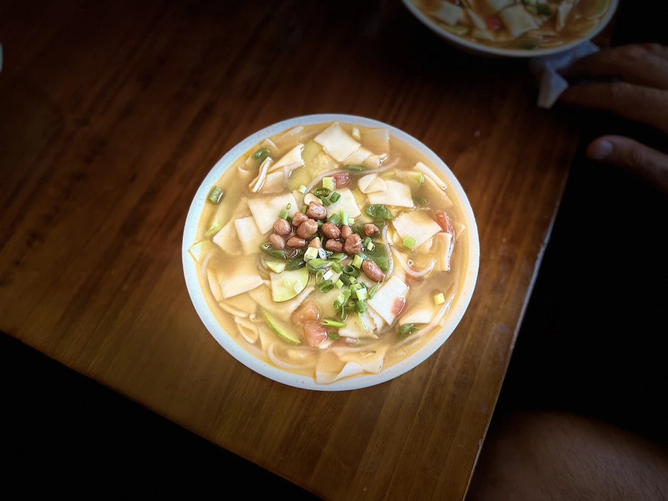 A top-down view of a bowl of traditional thenthuk noodle soup with hand-pulled noodles and fresh greens, representing authentic Tibetan food on a wooden table.