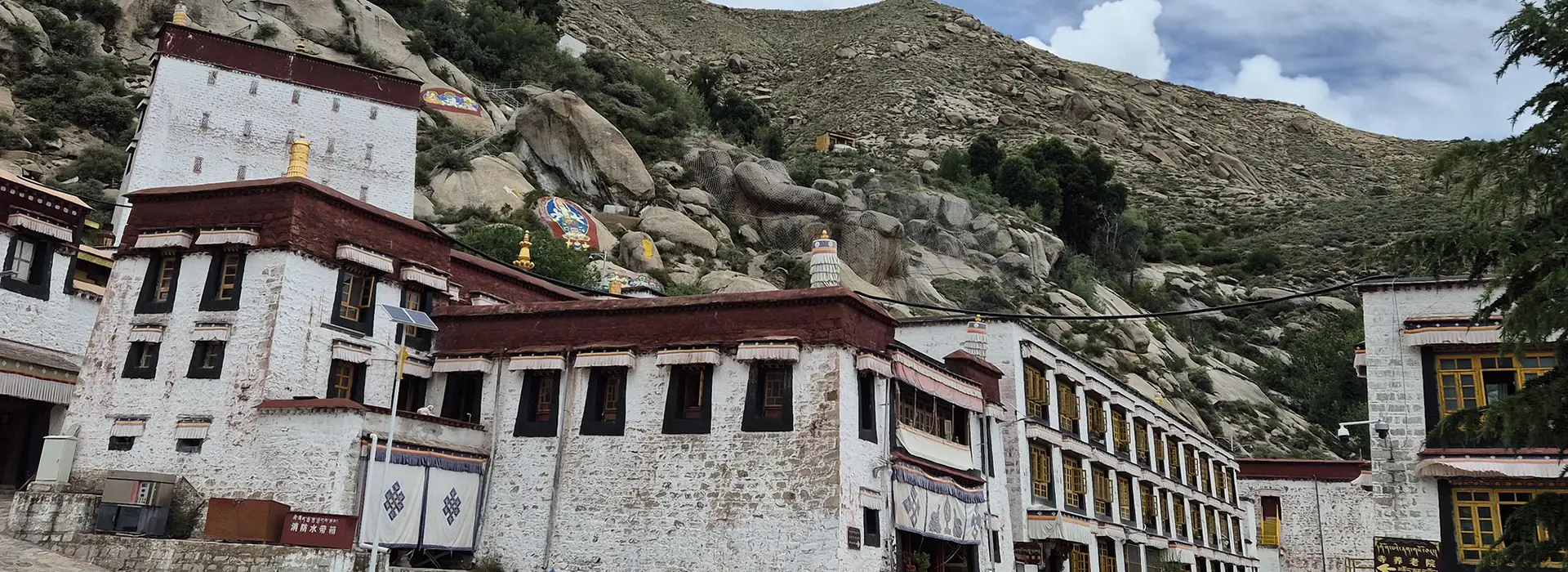 Traditional white and red stone monastic buildings sit nestled against a rocky hillside under a bright sky during a Lhasa and Yamdrok Lake Tour.