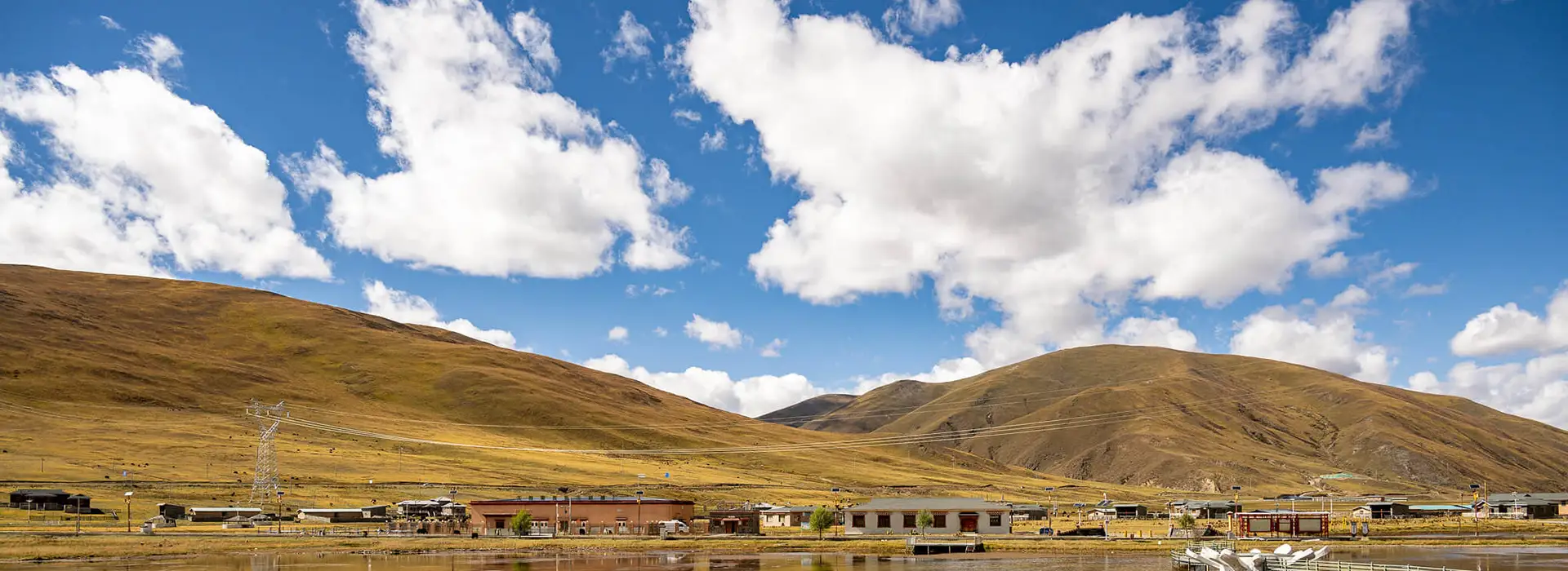 The calm waters of a high-altitude lake reflect a remote village and golden hills under a cloudy sky, requiring items from your Tibet packing list.