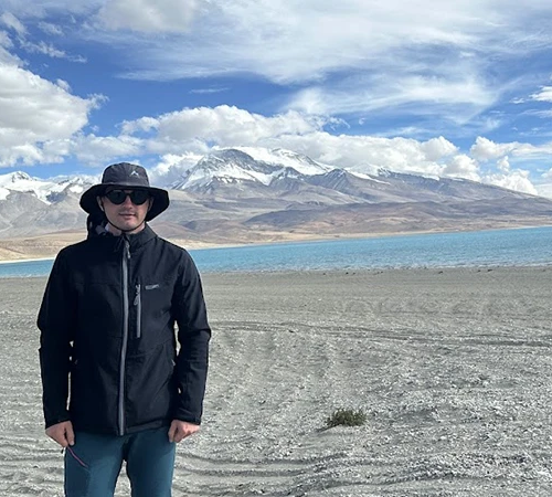 A male traveler wears sunglasses and a hat by a bright blue Tibetan lake. He secured his entry permit through the specific Tibet visa process in Kathmandu.