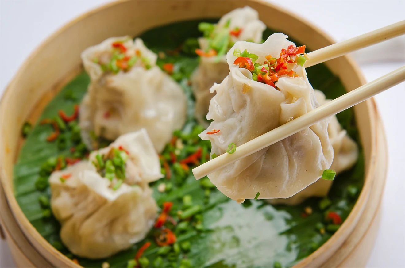 Chopsticks lift a steamed momo dumpling topped with chili and green onions from a bamboo steamer, showcasing a famous staple of Tibetan food.