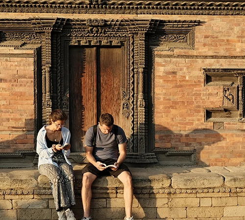 Two travelers sit on a stone wall in front of an ancient carved wooden door in Nepal, referencing their Nepal Travel Budget Guide for trip planning.