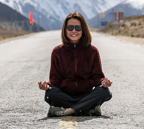 A traveler meditates cross-legged on a remote Tibetan road with snow-capped mountains and prayer flags in the background during a Lhasa and Shigatse tour.