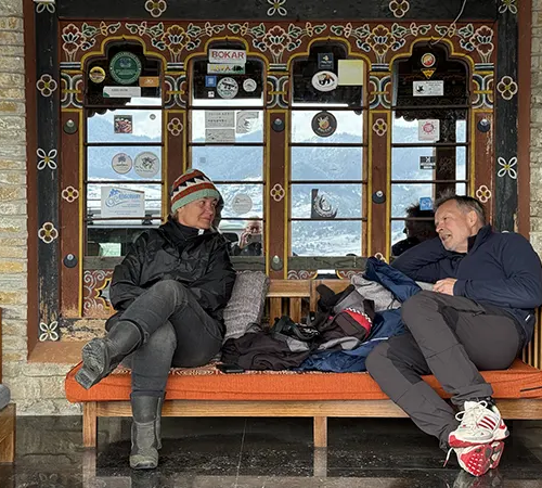 Two travelers relax on a wooden bench in front of a traditionally decorated window while enjoying a break during their Bumthang Cultural Trek and Tour.