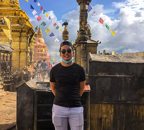 A smiling young traveler stands at a historic Newari temple in Patan, one of the best cultural destinations in Nepal, holding a phone with distant travelers.