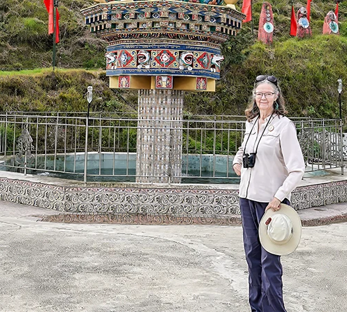 A smiling woman with binoculars and a sun hat poses near a colorful, traditional water prayer wheel during her immersive Western Bhutan Exploration.