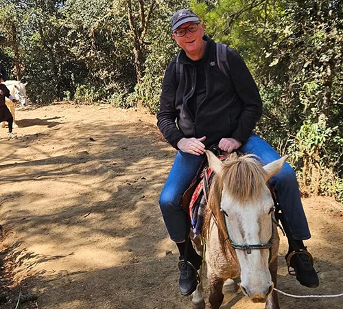 A traveler rides a light-colored horse on a dusty mountain trail during a Phobjikha Valley Nature Tour. Lush green trees and other riders appear in the background.