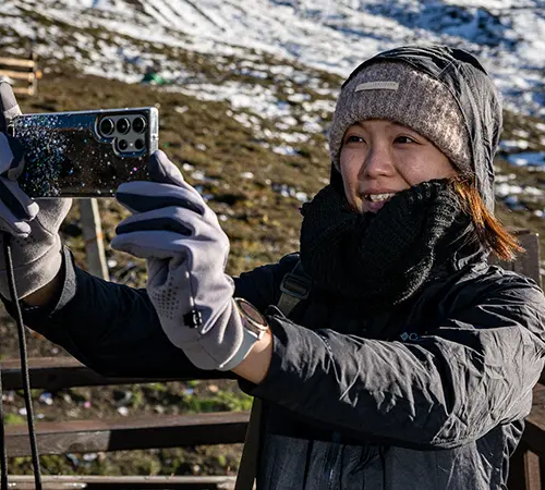 A smiling female traveler in warm, layered clothing, wearing a wool beanie and grey gloves, takes a high-altitude selfie. Exploring these sensitive regions requires obtaining the essential Alien's Travel Permit.