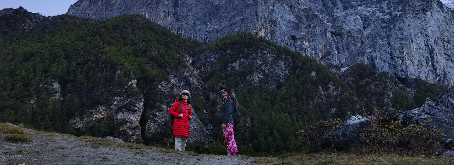 Two female travelers in winter jackets stand before a massive, jagged mountain cliff during a guided Bhutan Botanical Tour through the high Himalayas.