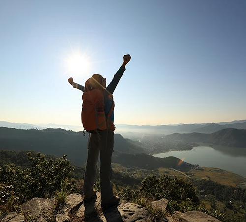 A hiker with an orange backpack cheers atop a hill overlooking Phewa Lake in Pokhara. Reviewing Nepal Travel Safety Tips ensures a joyful and secure adventure.