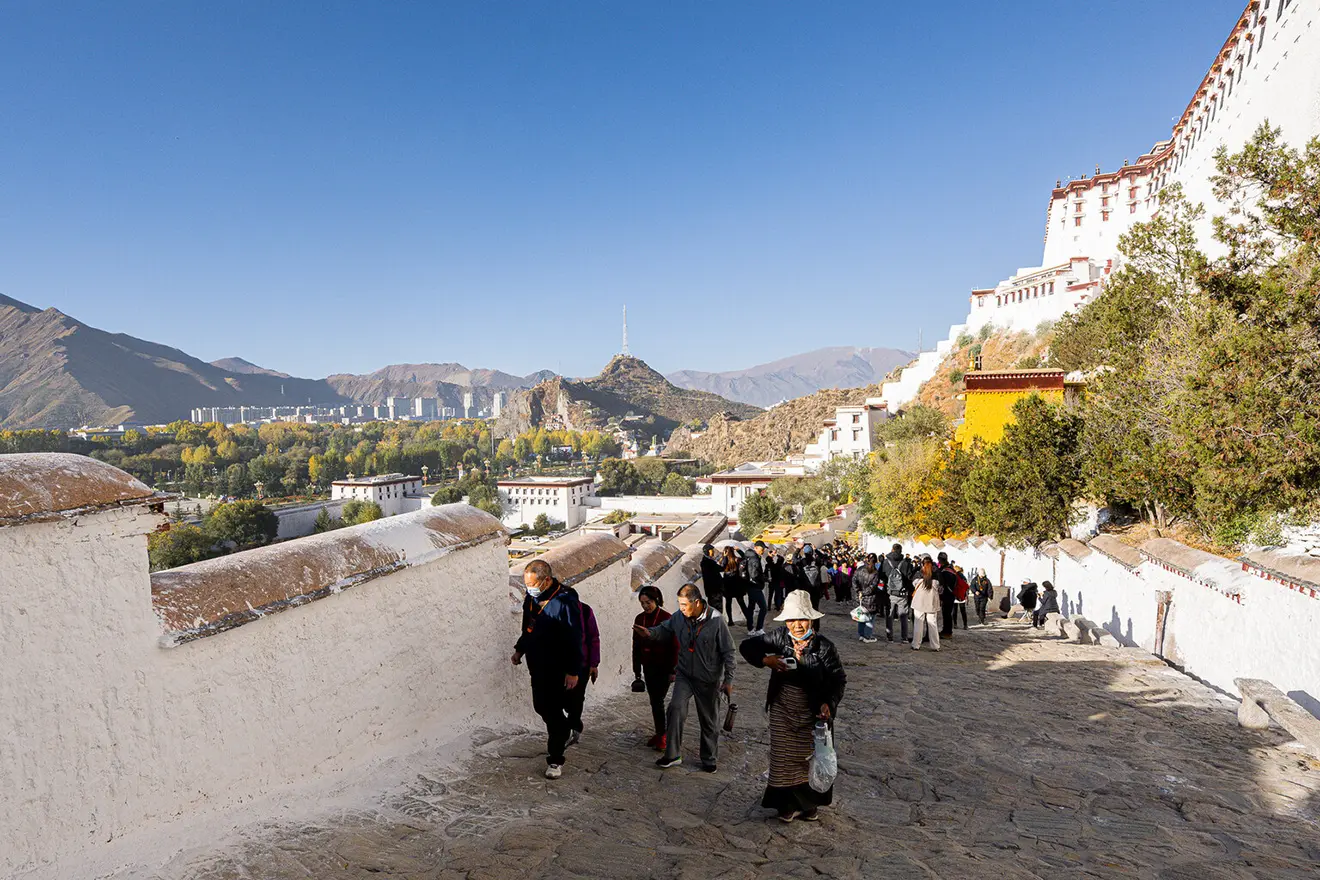 A crowd of people ascends the white-walled stone staircase leading up to the historic Potala Palace. Understanding Tibet Tour Requirements is essential before visiting this sacred, high-altitude site.