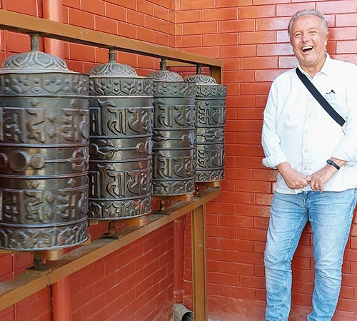 A cheerful traveler in a white shirt stands next to large prayer wheels in Kathmandu. Engaging with local spiritual customs is a top entry on any Nepal Travel Checklist.