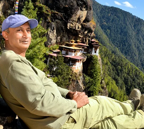A traveler with a backpack sits cross legged on a high rock, looking across a pine valley towards Tiger's Nest Monastery on a Bhutan Highlights Tour.