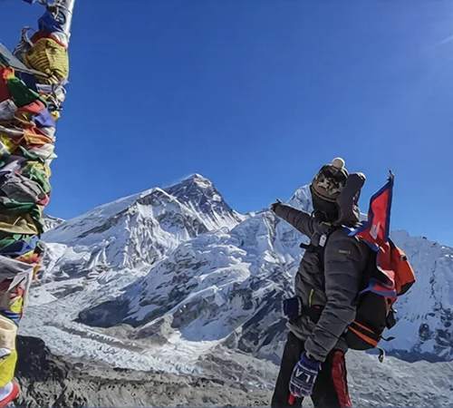 A hiker in winter gear points toward the snowy summit of Mount Everest near colorful prayer flags, capturing a triumphant moment on the Everest Three Passes Trek.