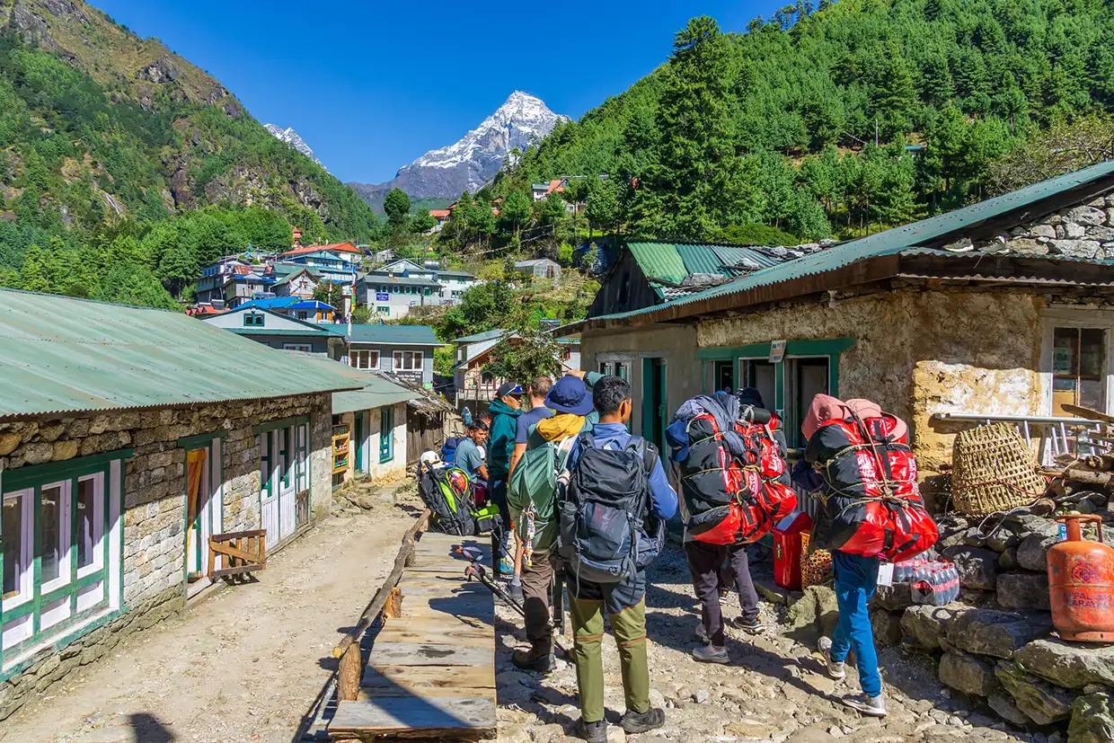 Trekkers with large backpacks walk through a sunlit mountain village toward a snow-capped peak, highlighting the gear found on a professional Nepal Hiking Packing List.