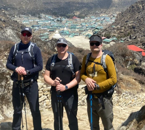 Three hikers with trekking poles stand on a mountain trail overlooking a village. They use professional gear to plan a trip to Nepal safely.