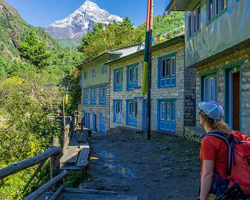 A hiker with a red backpack gazes at a snow-covered mountain peak from a stone village path, illustrating the value of a Nepal Hiking Permits Guide.
