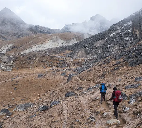 Two trekkers with blue and red backpacks hike along a narrow dirt trail through a rugged, rocky Himalayan valley, following a professional Gokyo Lakes Trek Guide.