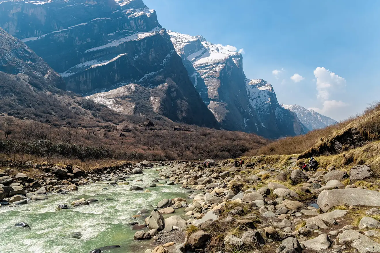 A rocky river flows through a deep valley toward massive snow-dusted cliffs, capturing the rugged terrain found in the best mountain destinations in Nepal.