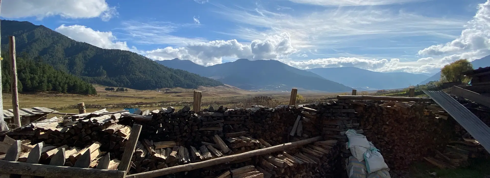 A scenic view of Bhutan's Phobjikha Valley from behind large stacks of drying firewood, perfect for a Phobjikha Valley Nature Tour.