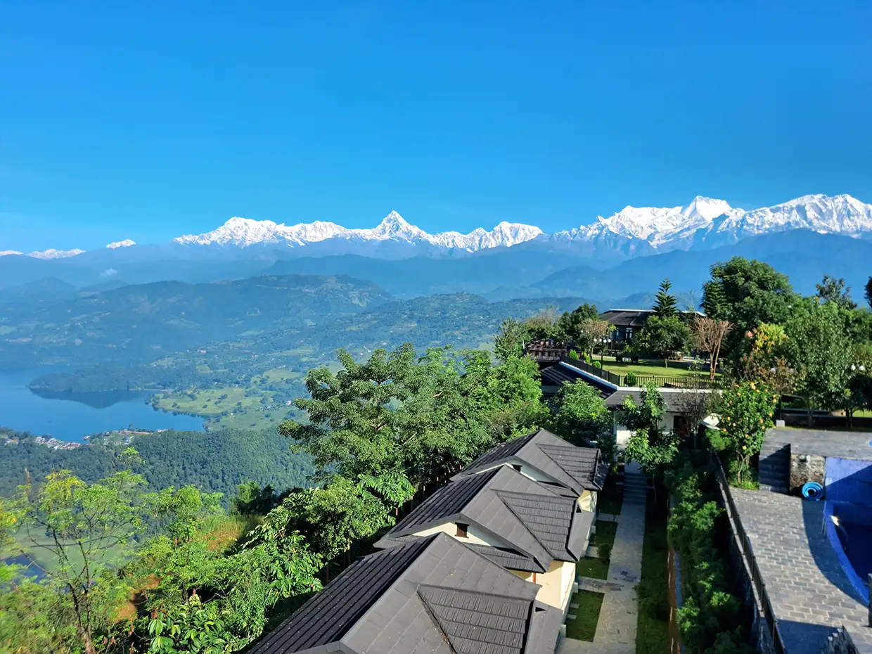 A scenic high-angle view captures a luxury resort with traditional roofs and a pool overlooking the clear blue Phewa Lake and the snow-capped Himalayan peaks, as detailed in our comprehensive Nepal Travel Budget Guide.