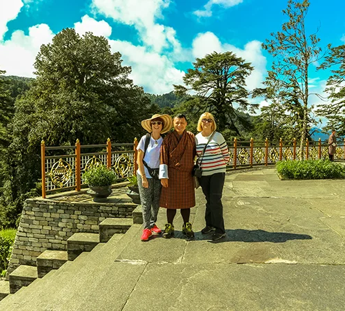 Two female travelers pose with their local guide on a stone terrace overlooking lush pine forests during a Classic Bhutan Tour.