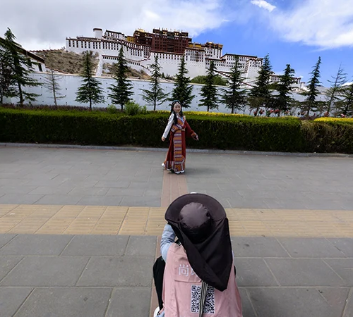 A person in a pink safety vest takes a picture of a girl in traditional Tibetan clothing in front of the Potala Palace during a Lhasa to Everest Base Camp and Namtso Lake Tour.