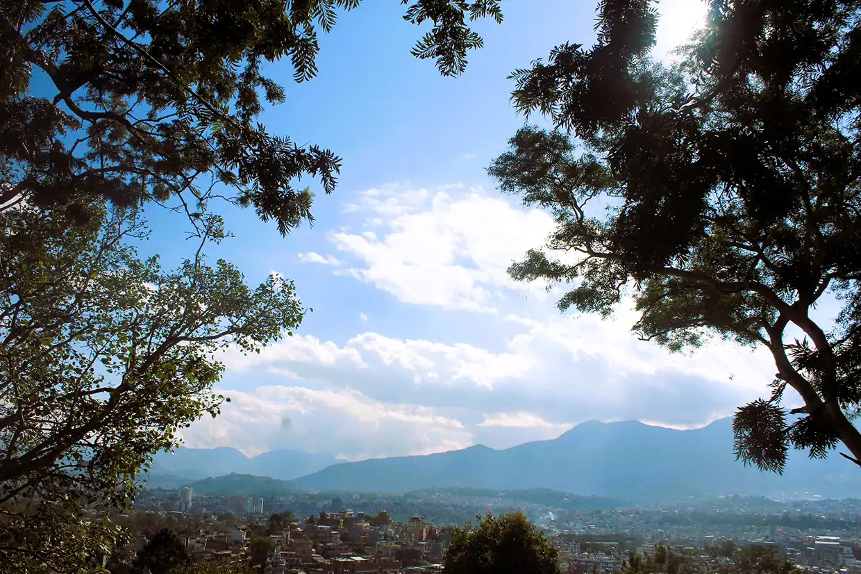 A wide view of the city valley and distant mountains framed by green trees under a bright blue sky. This panoramic vista is among the top things to do in Kathmandu.