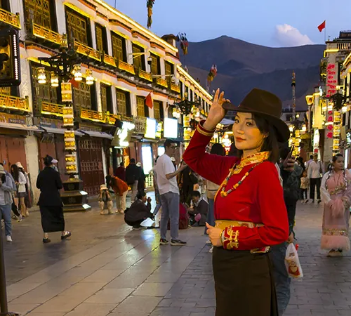 A traveler in traditional attire adjusts her hat on a brightly lit street in Lhasa at dusk, captured during a Lhasa Short Tour.