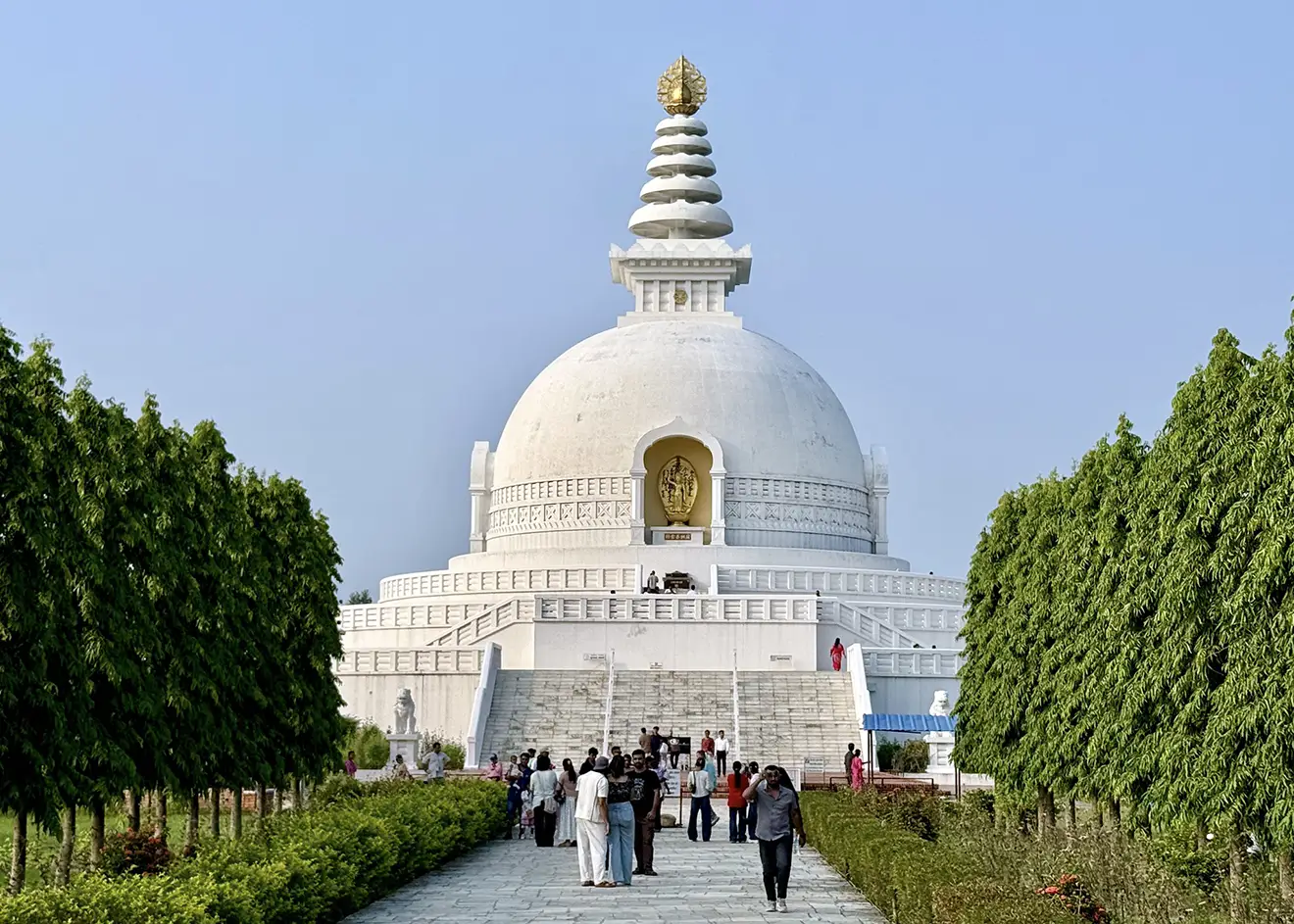 The large, white World Peace Pagoda stupa with a gold finial rises above a line of green hedges and walking paths, a key spiritual stop during a 10-Day Nepal Itinerary.
