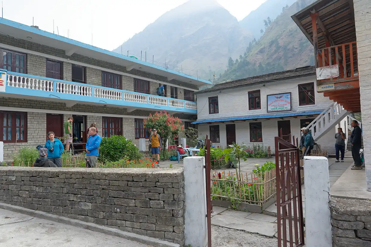 Trekkers relax in the courtyard garden of a stone guest house surrounded by steep mountain peaks during the Nar Phu Valley with Annapurna Circuit Trek.