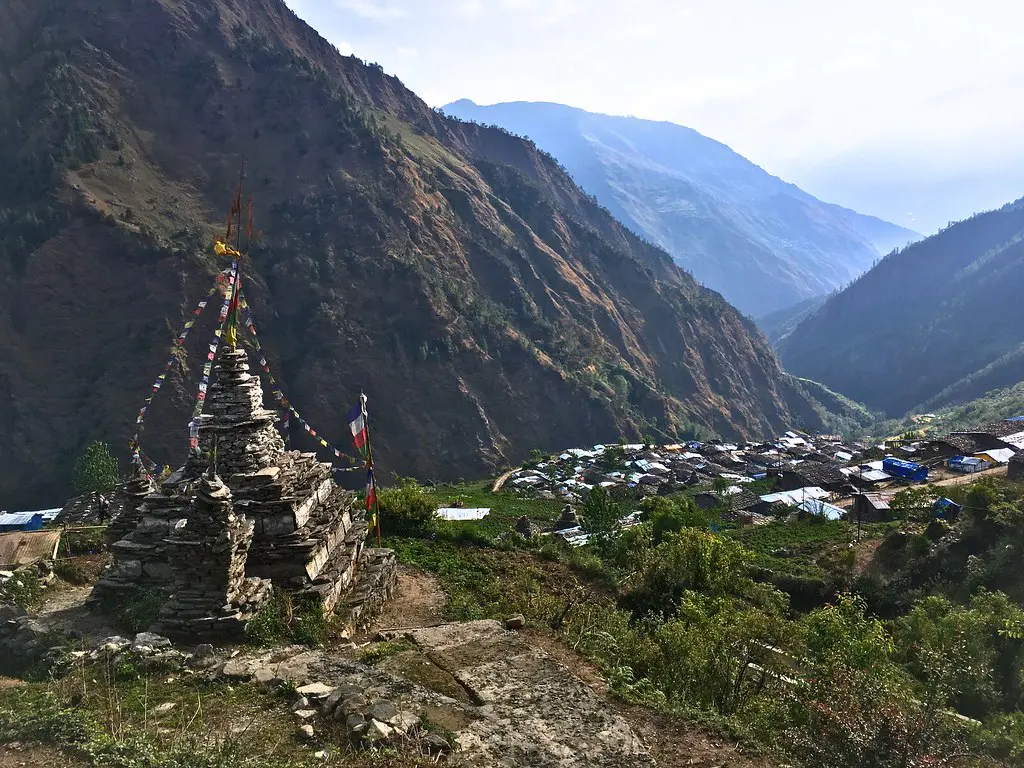 A stone chorten draped in colorful prayer flags overlooks a hillside village nestled in a deep mountain valley on a Tamang Heritage Trek.