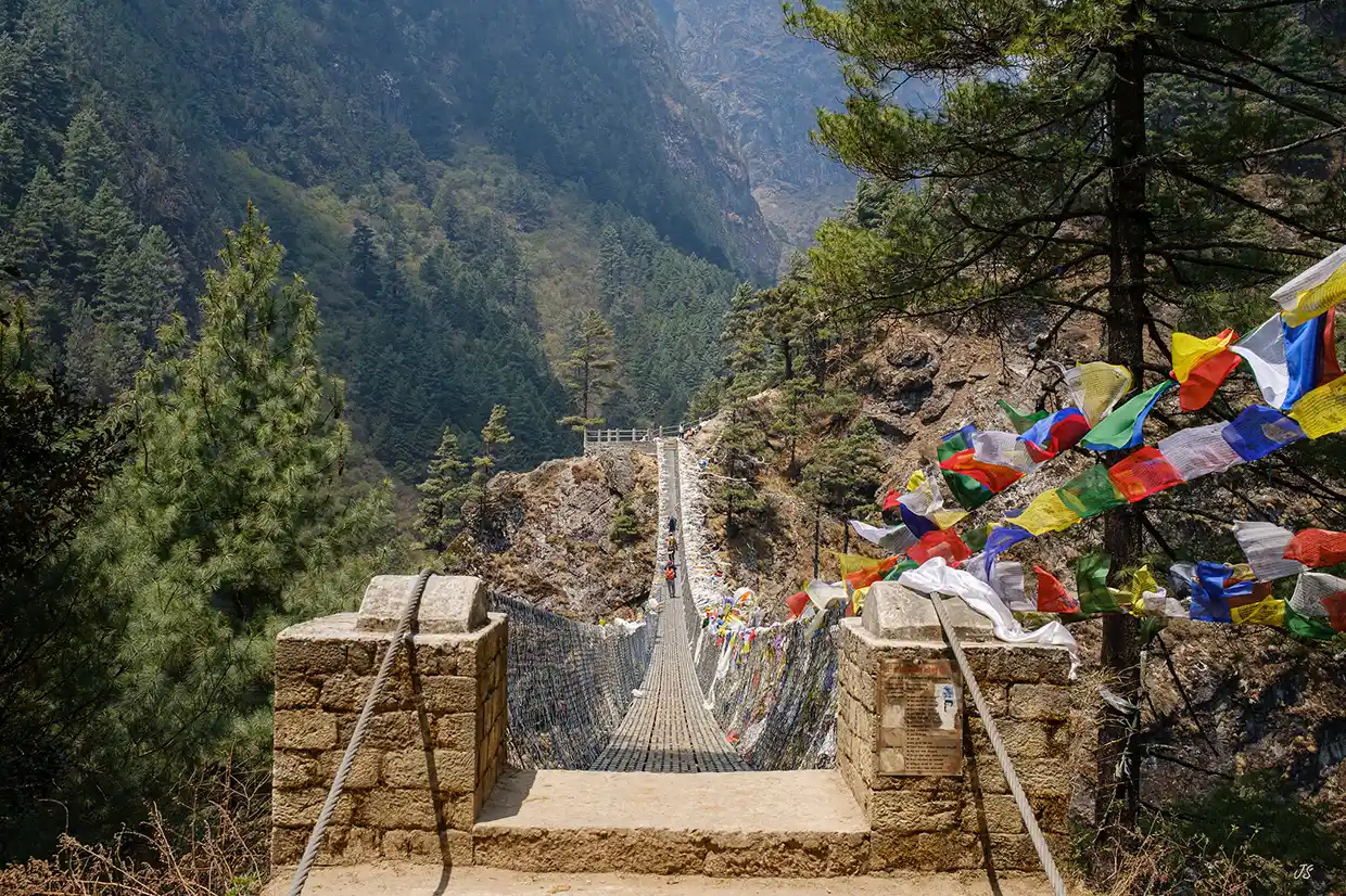 The high Hillary Suspension Bridge stretches across a deep, forested valley filled with colorful prayer flags, welcoming hikers on an Everest Cultural Trek.