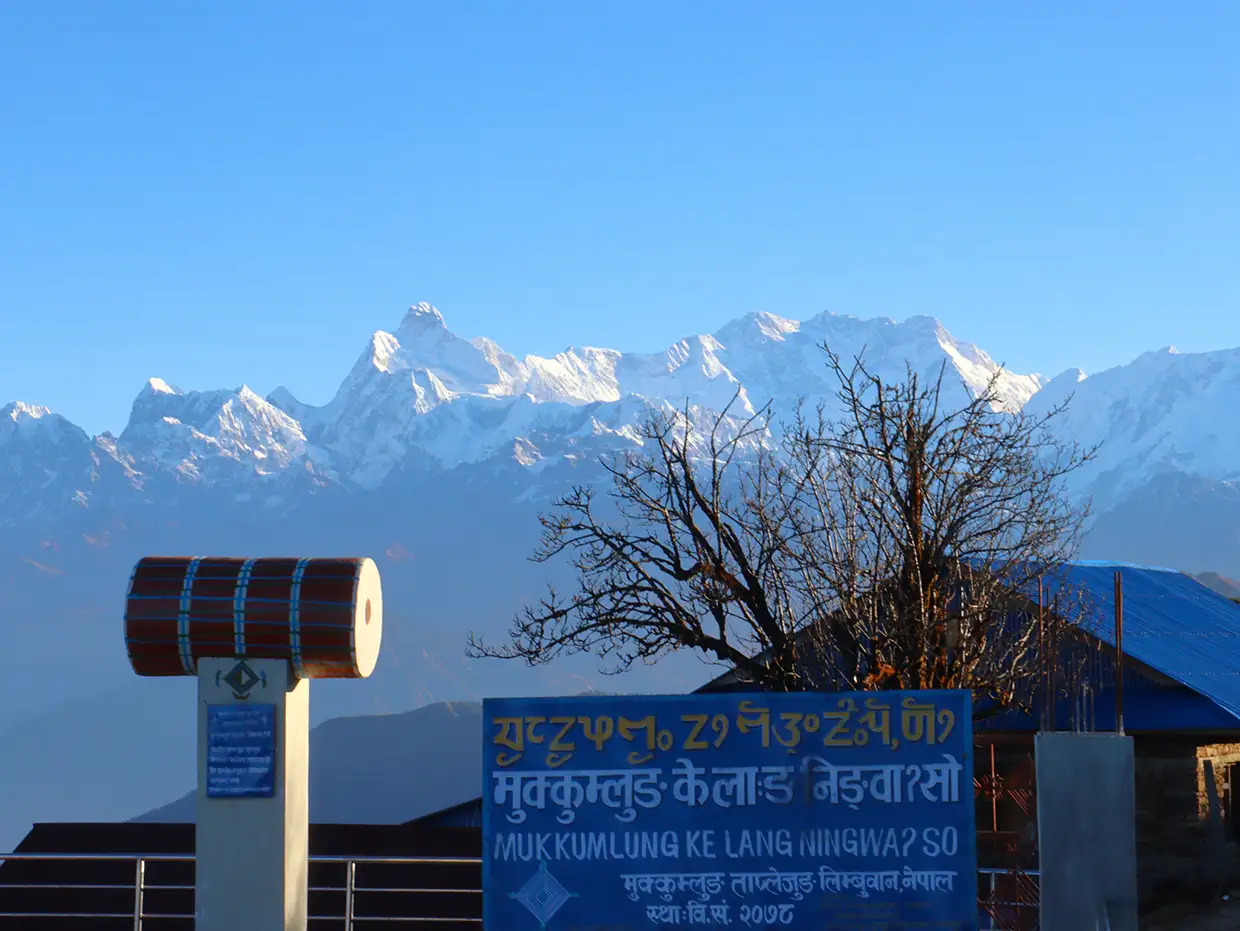A clear view of snow-capped Kanchenjunga mountains from Taplejung behind a local blue sign. Always check current Nepal Trekking Regulations before visiting this remote eastern region.