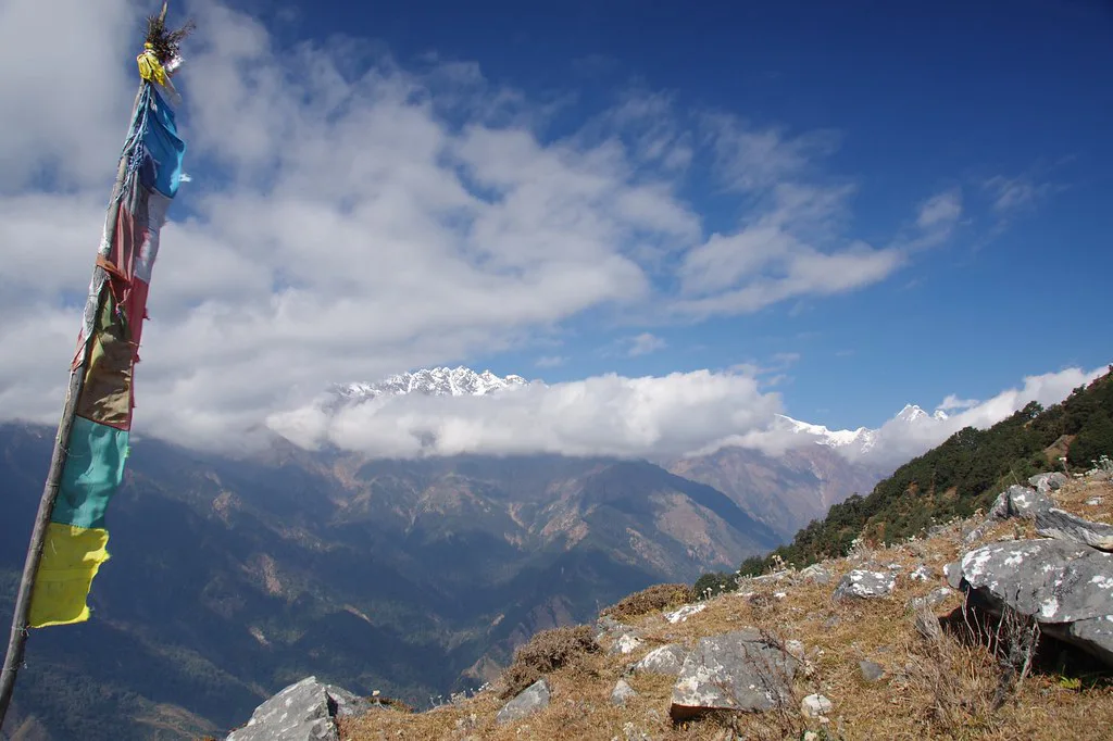 A colorful vertical prayer flag stands on a rocky mountain ridge overlooking snow-capped peaks and deep valleys under a partly cloudy sky on a Tamang Heritage Trek.