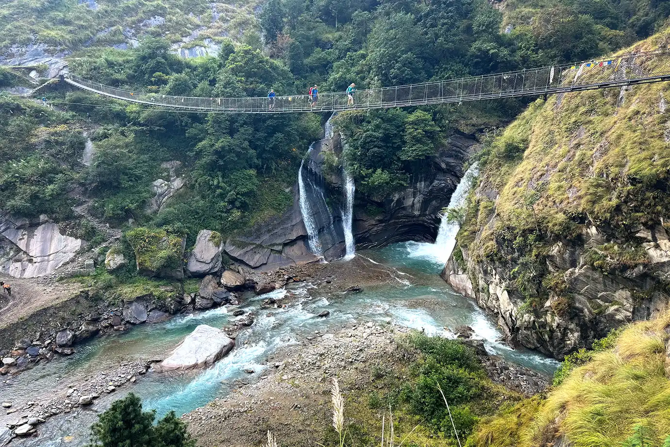 Trekkers cross a suspension bridge between Jagat and Deng on the Manaslu Circuit Trek, above a turquoise river and waterfalls.