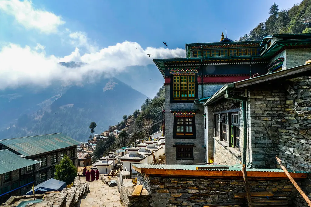 Buddhist monks walk down stone steps past the stone buildings of Thupten Choeling Monastery into a misty mountain valley during a Pikey Peak Trek.