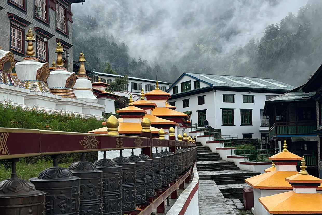 A row of bronze prayer wheels lines a stone path leading past white stupas at Thupten Choeling Monastery on a misty Pikey Peak Trek.